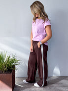 Person wearing a pink top and brown pants with white stripes, standing against a light gray wall.